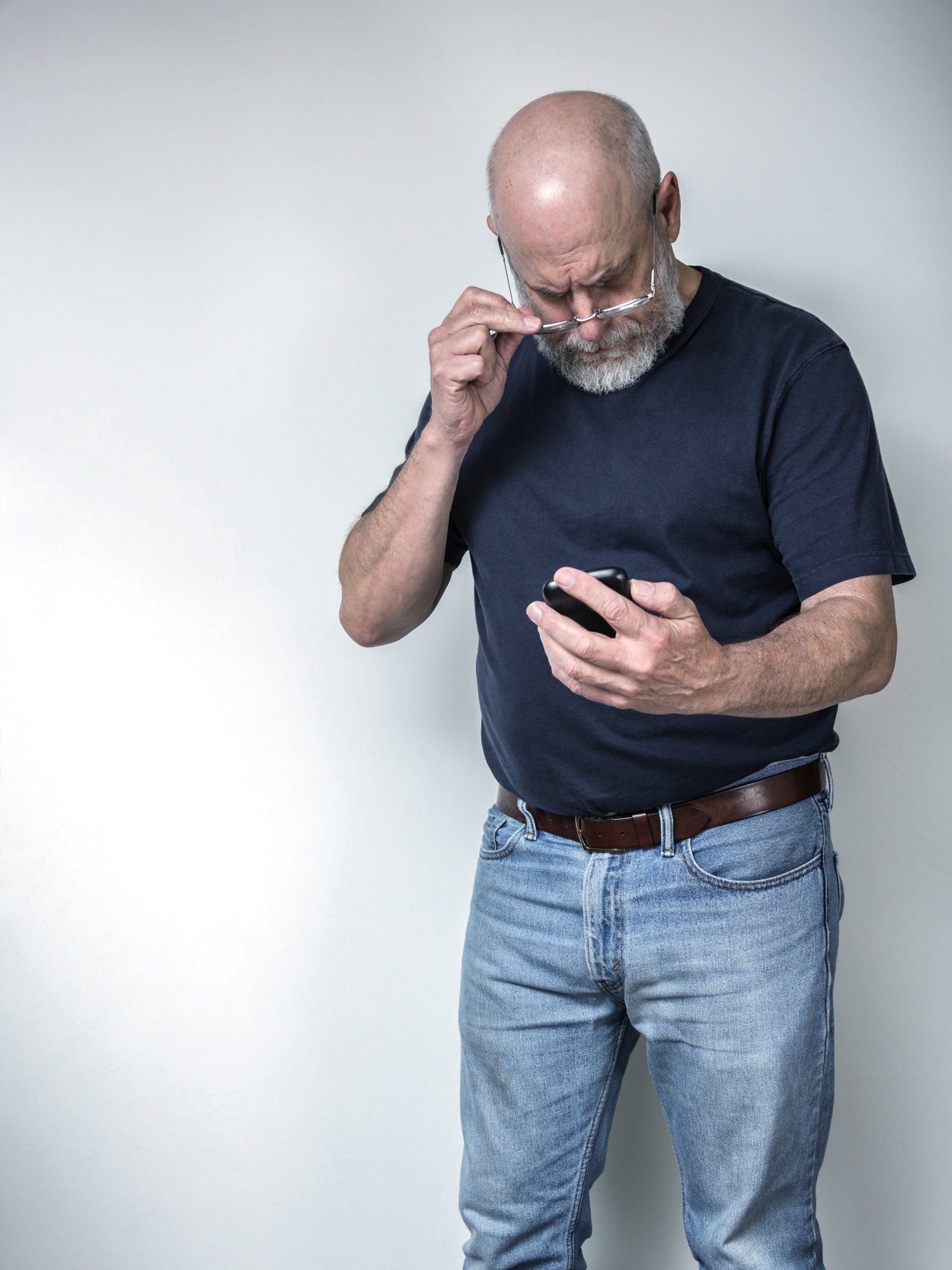 A senior adult man with poor eyesight is holding his eyeglasses away from his face as he looks down at the smartphone in his hands trying to read a text message. He is wearing a dark short sleeve t-shirt and blue jeans. He has a gray beard and mustache. Three quarter length on a grey background.